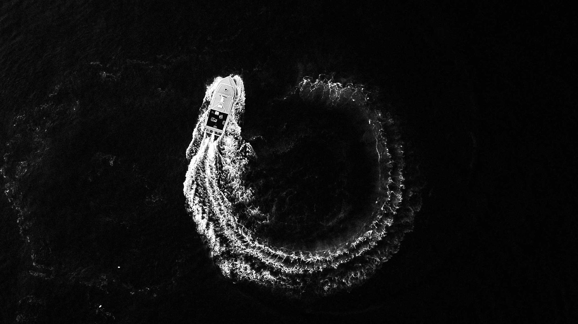 Aerial view of a boat carving a circle wake in deep black water.